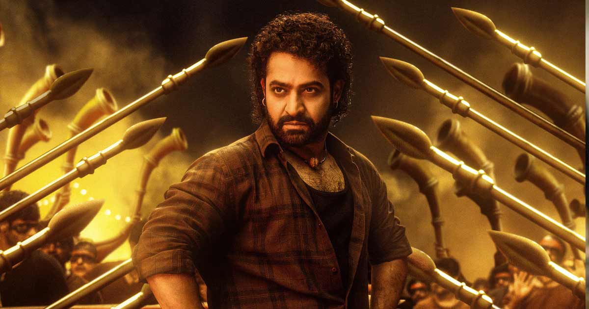 Promotional image from Devara: Part 1, an epic Indian action-drama film. The scene showcases lead actor Jr. NTR in intense battle armor, portraying a determined and heroic stance against a striking natural backdrop. The film's setting includes rugged landscapes, dramatic lighting, and traditional costumes, highlighting themes of bravery and sacrifice. Other characters stand by, symbolizing camaraderie and loyalty, with a vibrant and cinematic color palette that enhances the grandeur of the scene